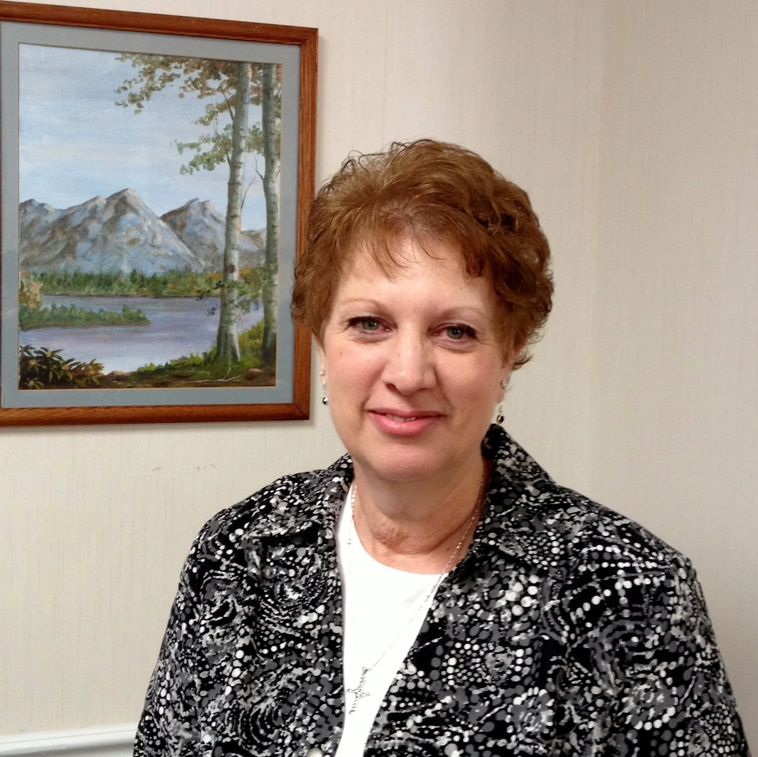 A middle-aged woman with short reddish-brown hair, wearing earrings and a black and white patterned blazer, smiling at the camera. She is standing in front of a framed landscape painting of mountains, trees, and a lake on a beige wall.