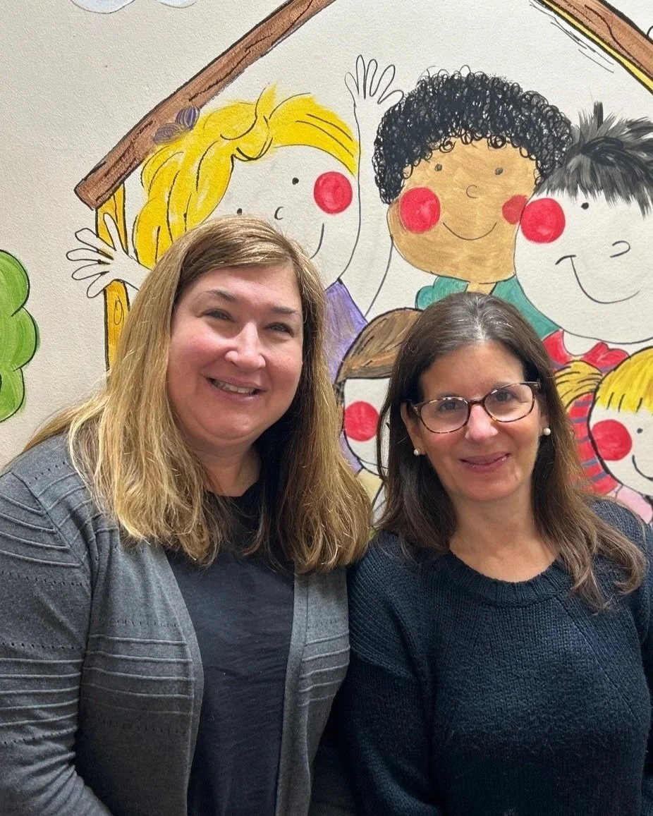 Two women standing in front of a colorful children's mural featuring cartoon children with big red noses and cheerful expressions.