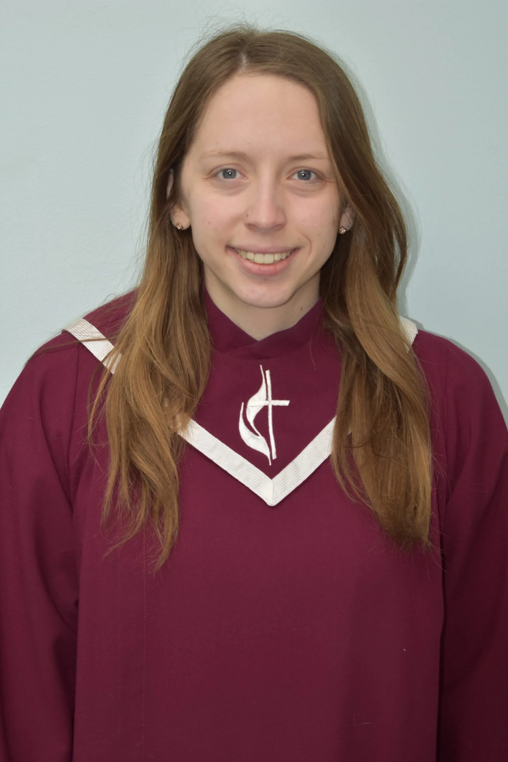 A young woman with long red hair, wearing a maroon choir robe with a white collar and a cross emblem, smiling in front of a plain light green wall.