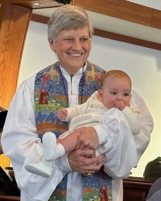 An older woman holding a baby in a church or religious setting. The woman is smiling, and the baby is making a facial expression with hands near the face, dressed in white. The woman is wearing a colorful vest with religious imagery.