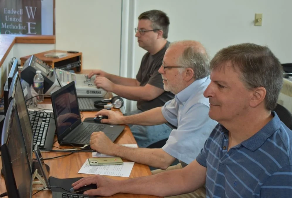 Three men working at a table with laptops, a mixer, headphones, and papers in a room with a whiteboard and a water bottle.