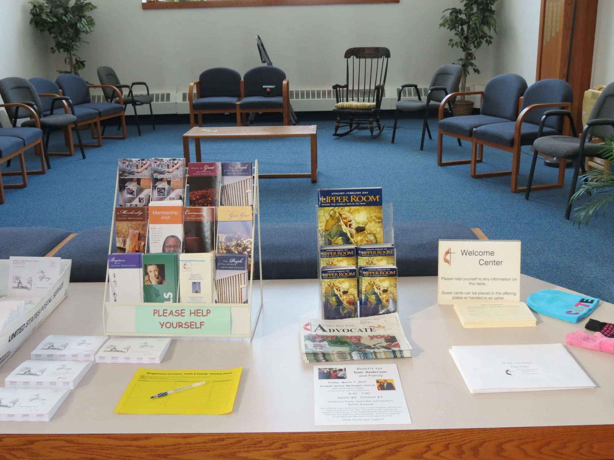Inside a church welcome center with chairs arranged in a semi-circle, informational brochures on a table, and a sign requesting donations and guest cards.
