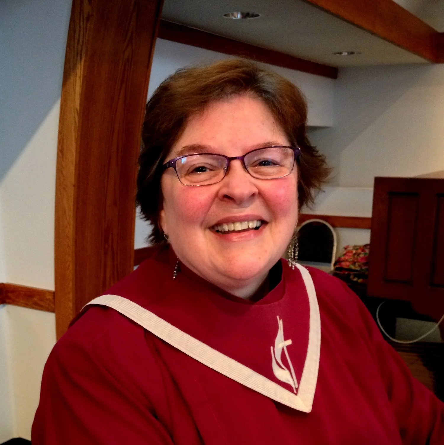 A smiling woman with short brown hair and glasses, wearing a red clerical shirt with a white collar and a cross necklace, standing indoors near a wooden beam.
