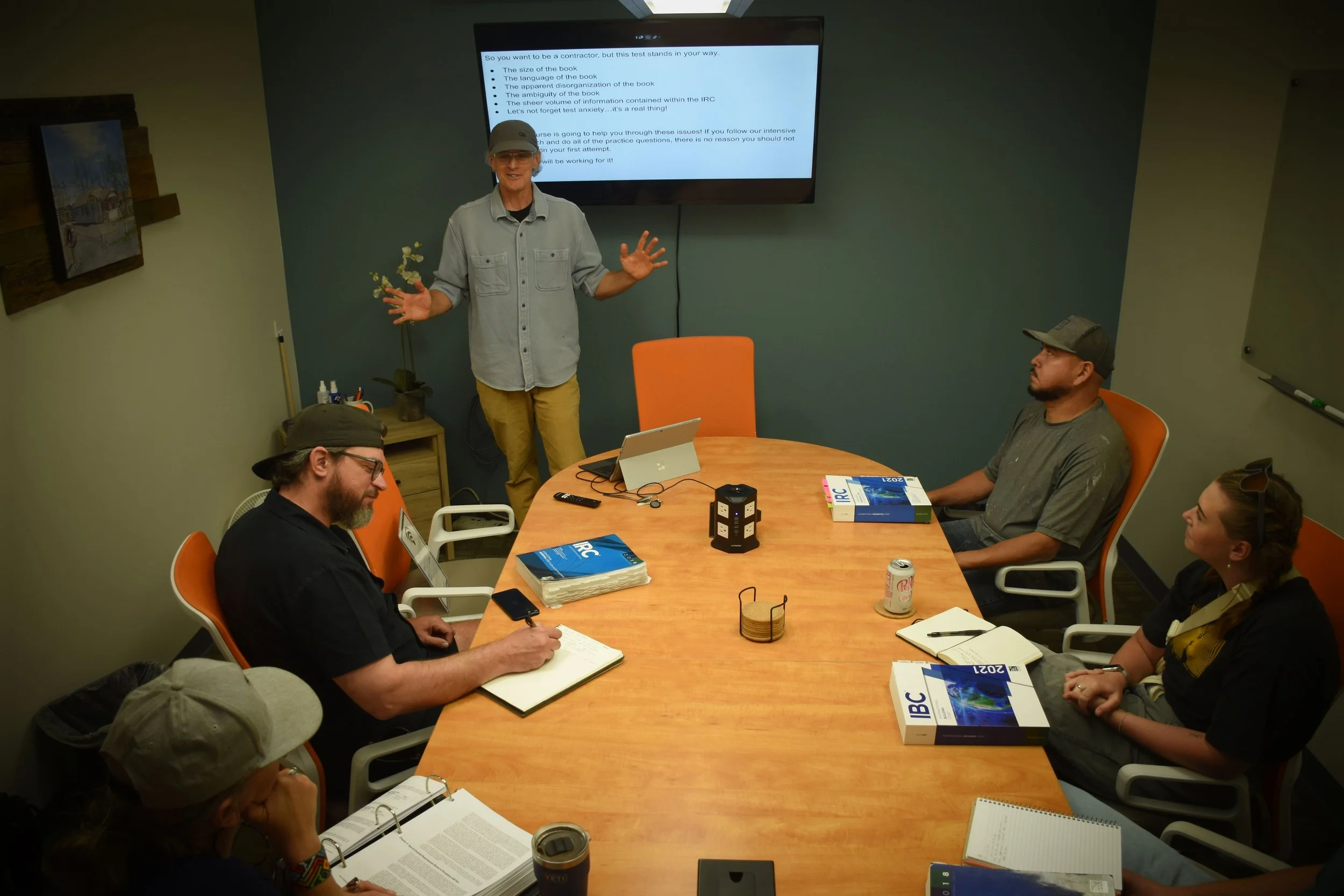 A man in a gray shirt and a baseball cap is standing and speaking in front of a room of seated people with books and notebooks, with a large presentation slide on the wall behind him.