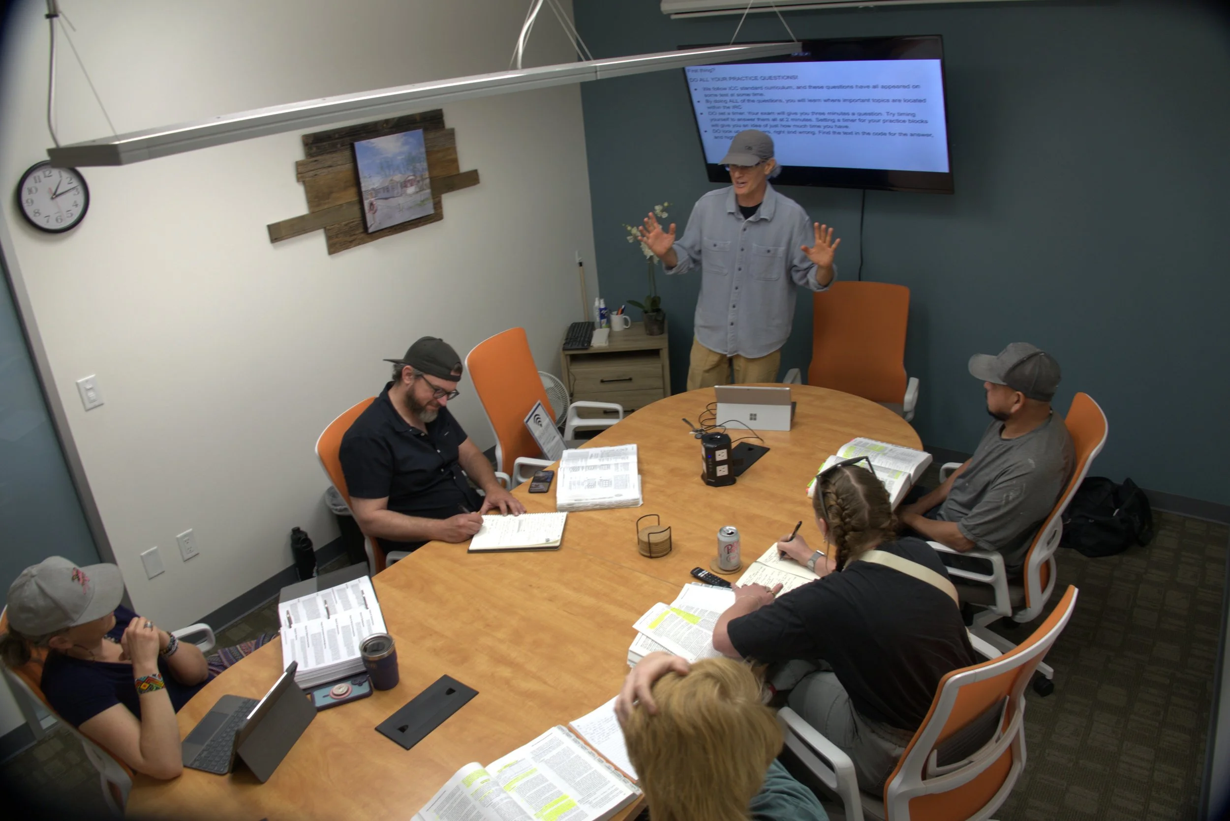 NCSI instructor teaching an in-person ICC G13 Residential Contractor exam prep class as students use the 2018 IRC code book, take notes, and practice code navigation from examples displayed on a large classroom screen.