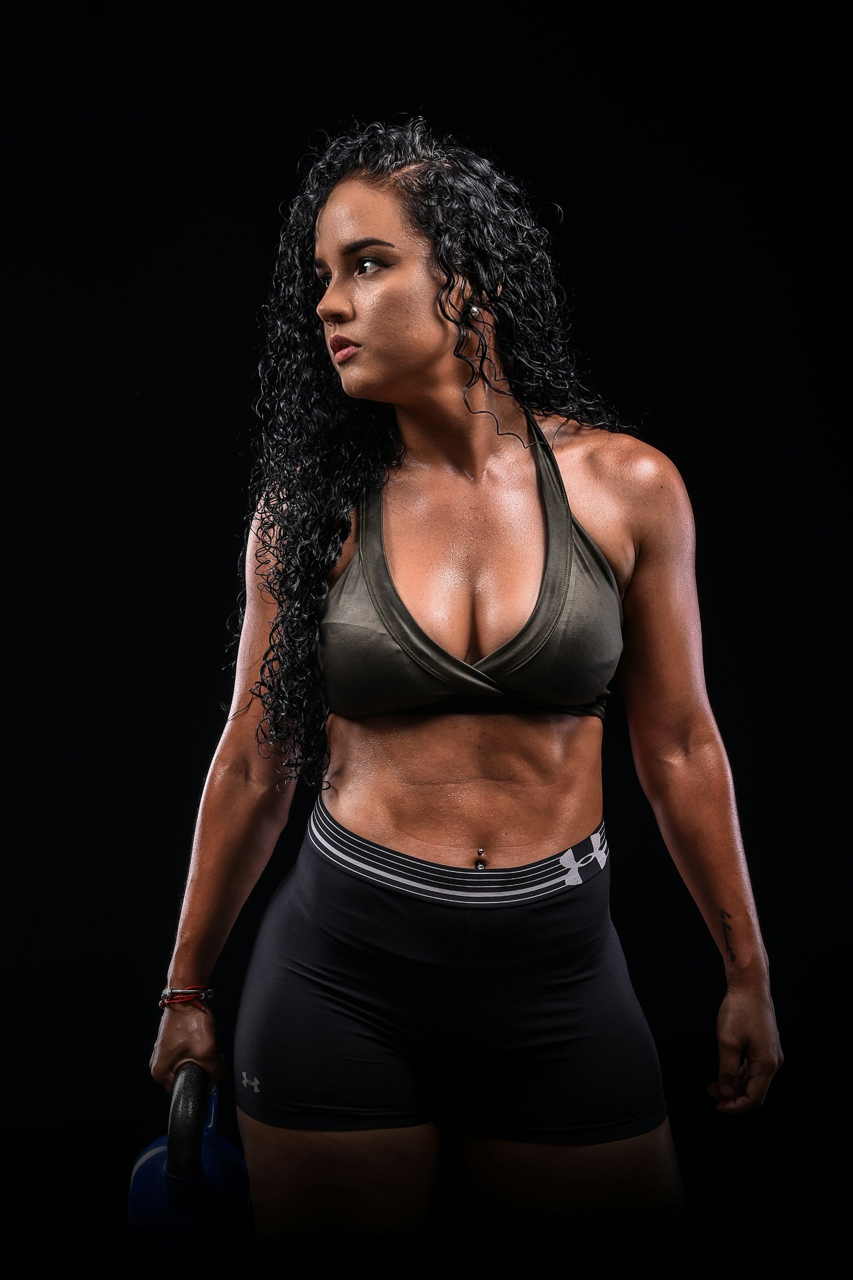 A woman with long, curly black hair wearing a dark sports bra and black workout pants standing against a black background. She is holding a kettlebell in her right hand, and her body appears sweaty, indicating she has been exercising.