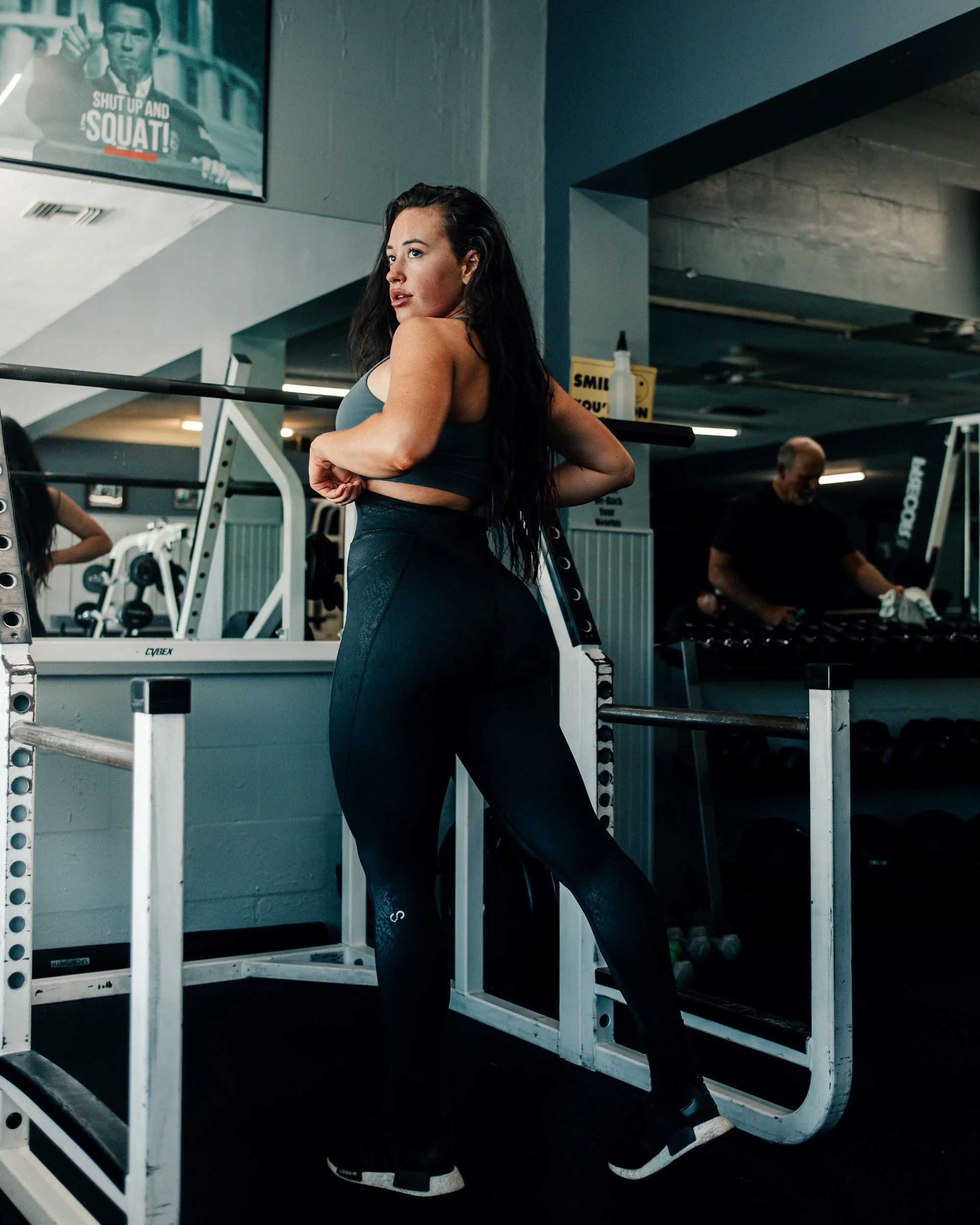 A woman in workout clothes standing in a gym, leaning on a squat rack, with a television screen showing a motivational message in the background.