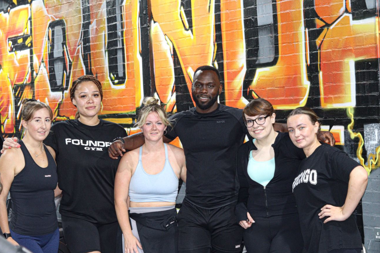 Group of six women and one man posing together in front of a colorful graffiti wall at a gym, with some members wearing workout clothes and smiling.