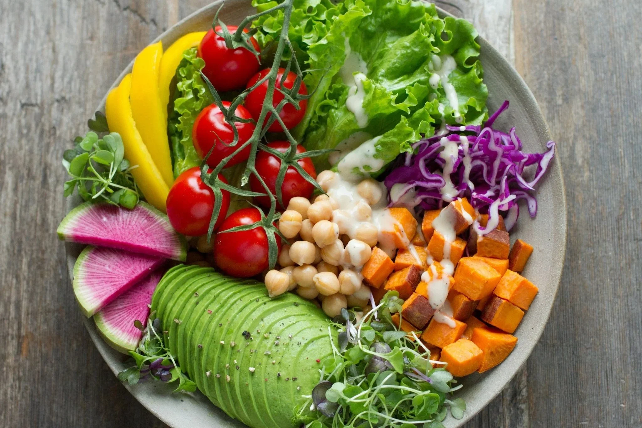 Assorted fresh vegetables and salad greens on a white plate, including cherry tomatoes, sliced radish, sliced avocado, chopped sweet potatoes, purple cabbage, shredded lettuce with dressing, and microgreens, on a rustic wooden surface.