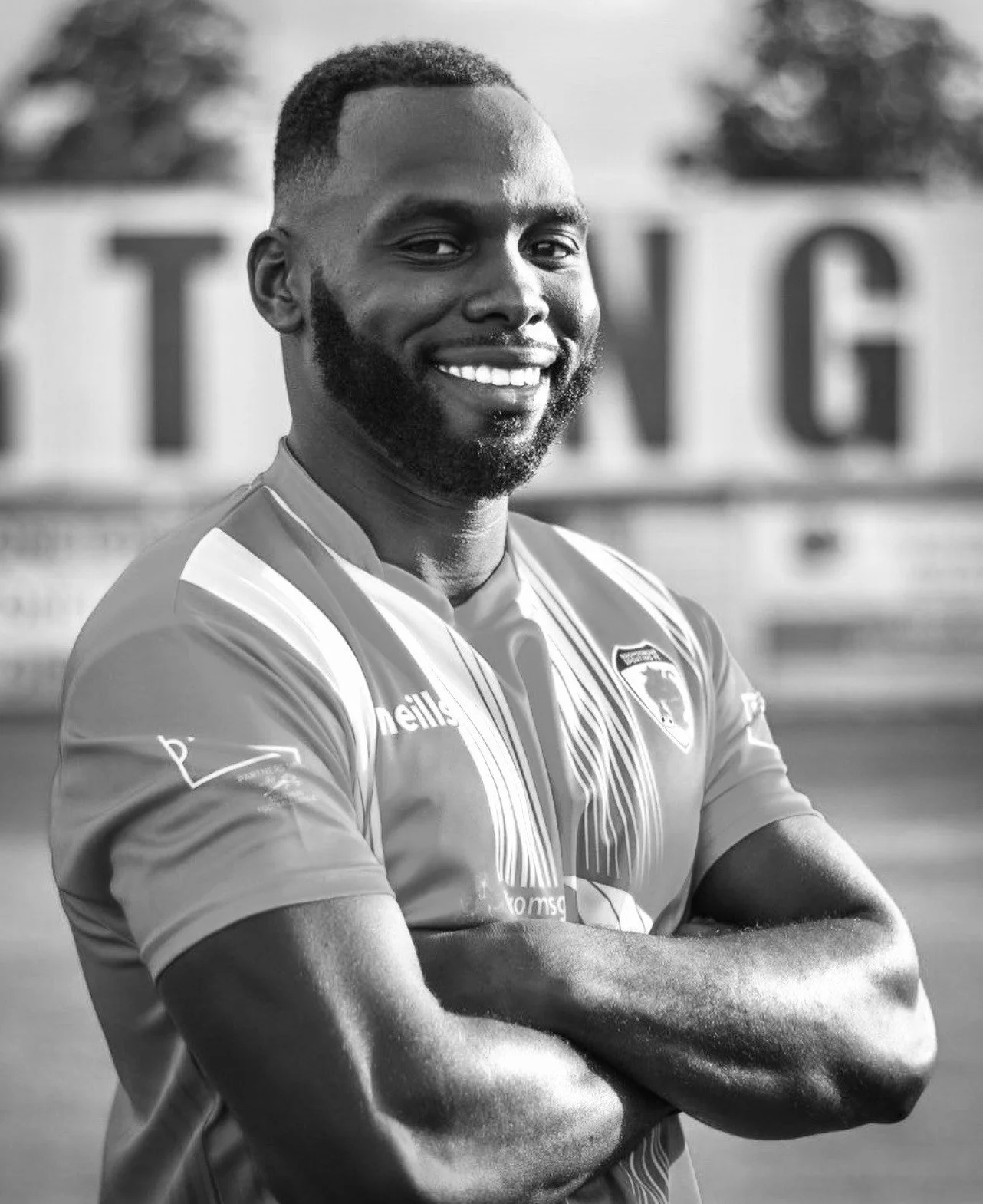 Black and white portrait of a smiling man with a beard and short hair wearing a sports jersey, standing outdoors with blurred background.