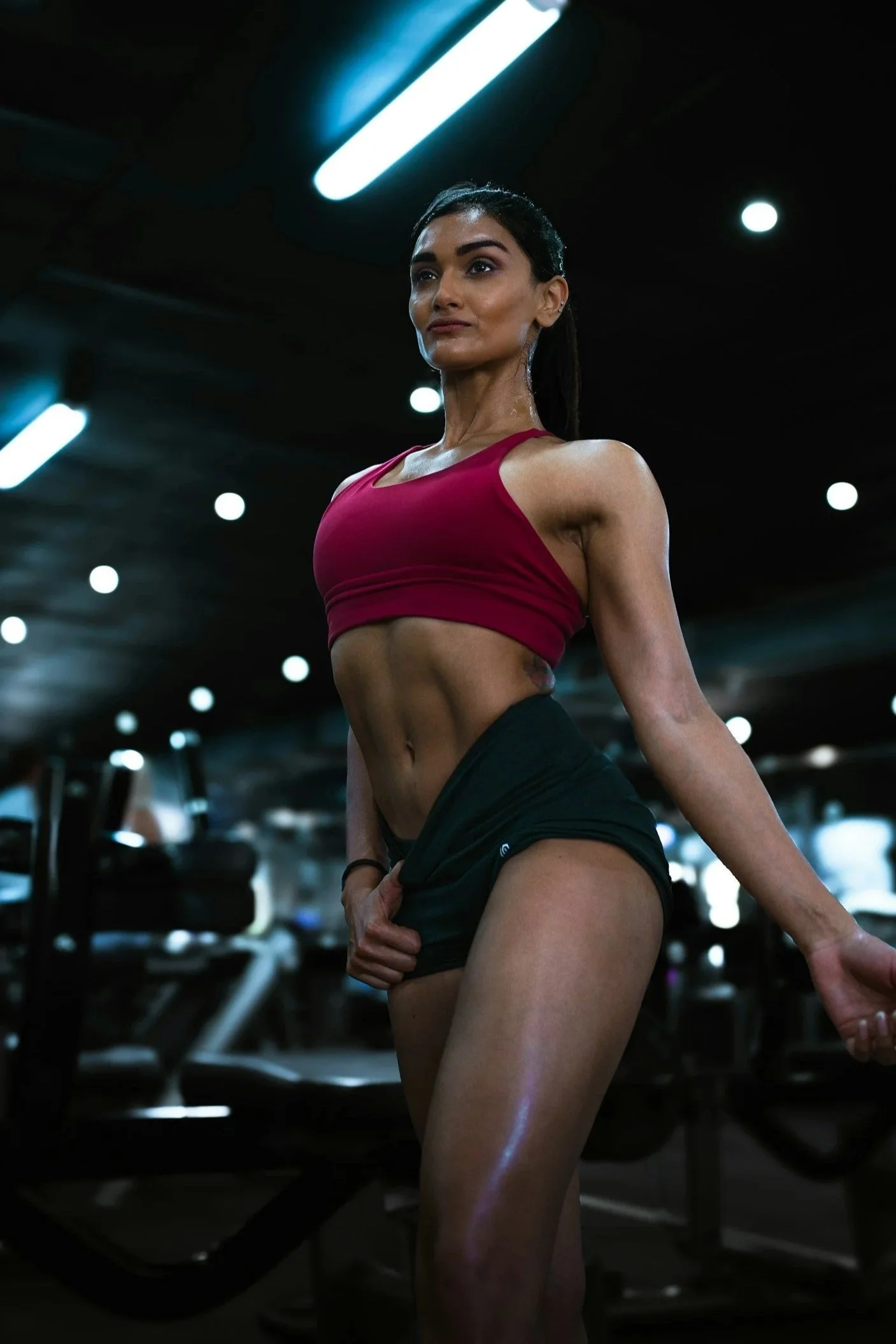 A fit woman in a burgundy sports bra and black shorts standing in a gym with black ceiling and bright overhead lights, touching her shorts and looking confidently to the side.