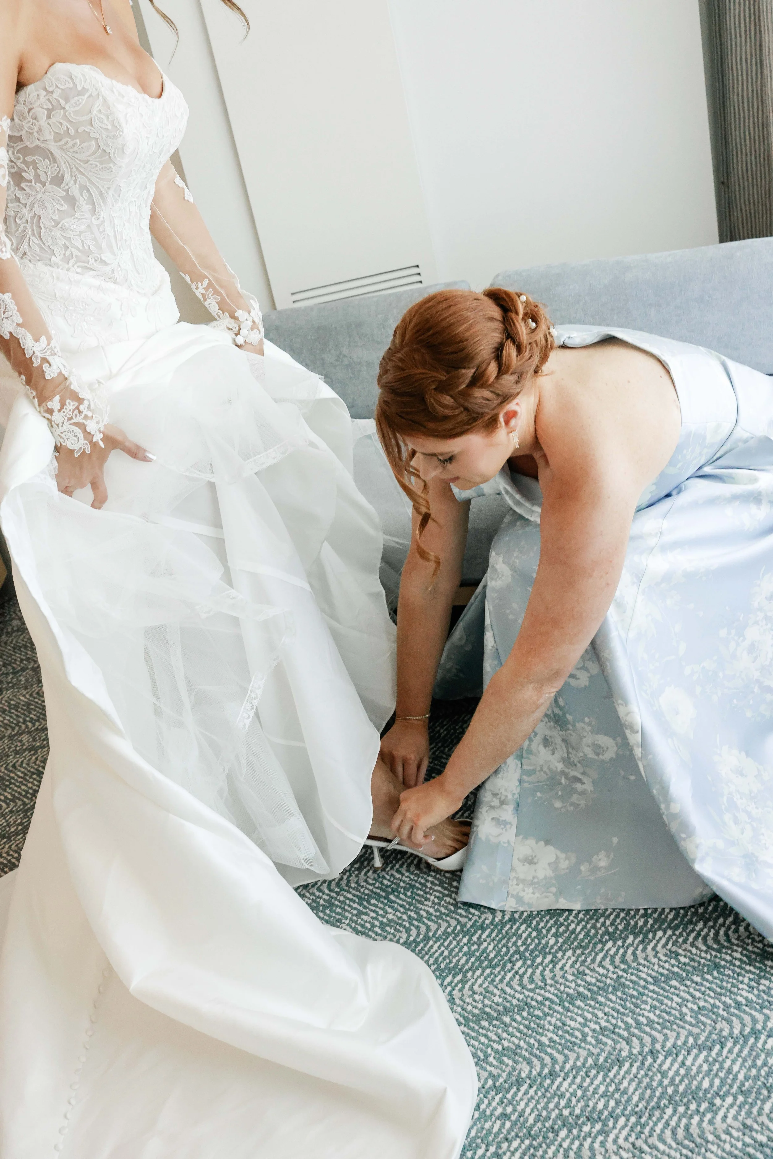 A bride wearing a lace wedding dress with long sheer sleeves is standing next to a woman in a blue floral dress who is kneeling and tying the bride's shoe.