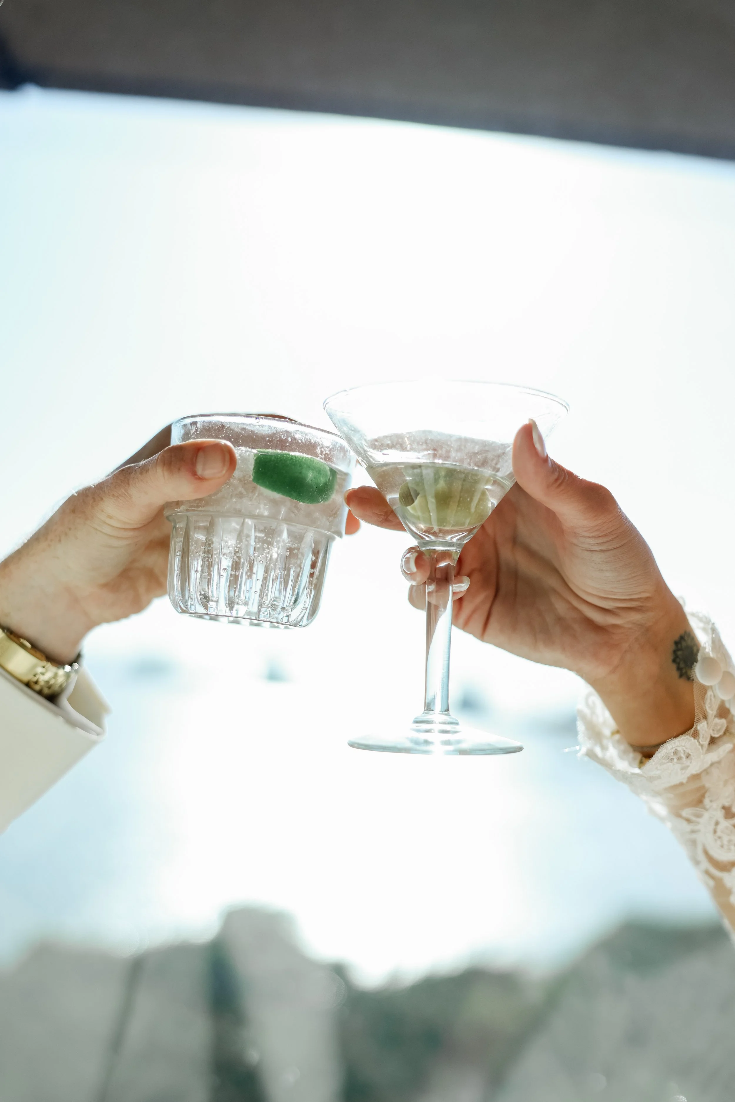 Two hands holding glasses with lime slices, clinking in a toast against a bright background.