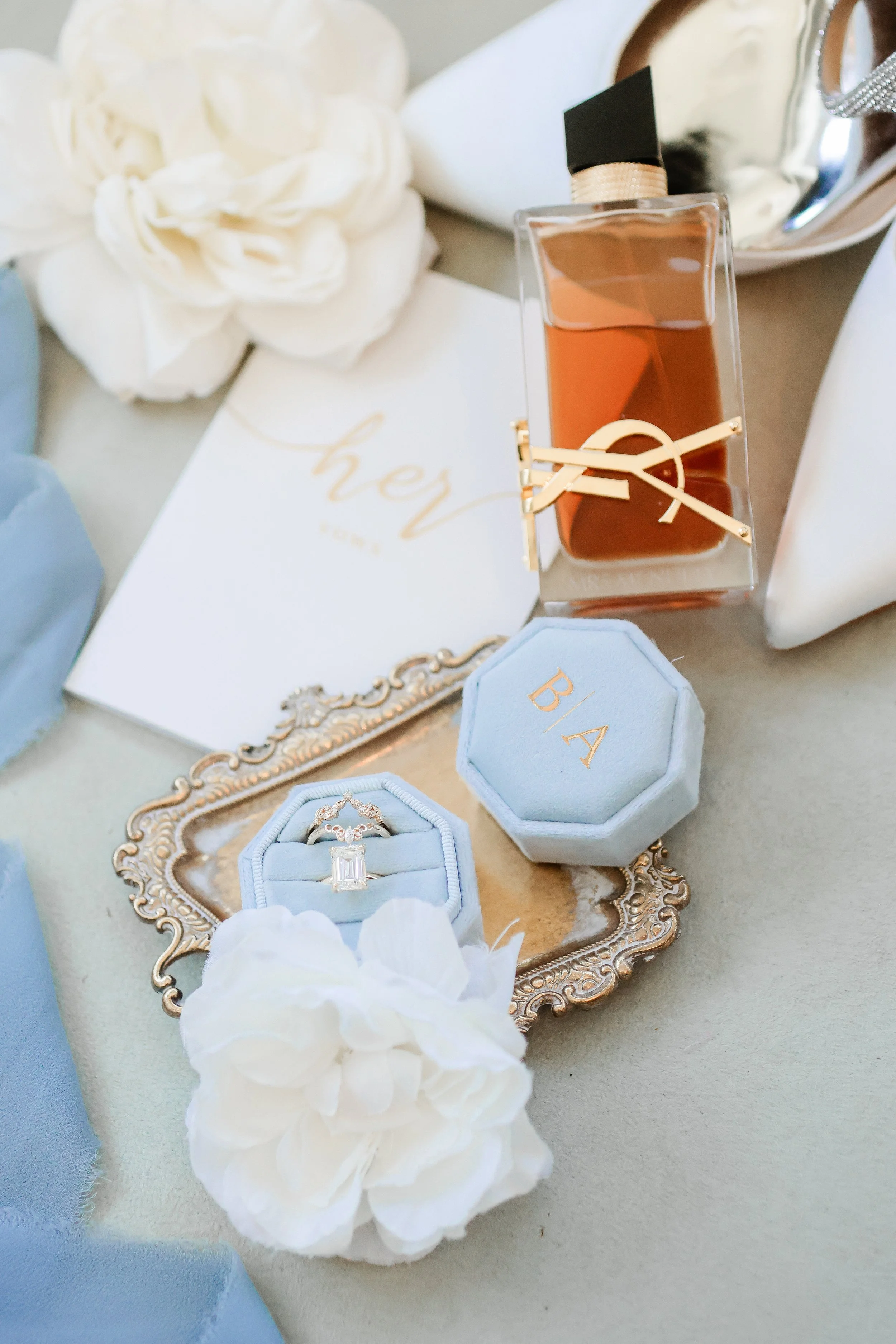 Wedding jewelry and perfume arranged on a decorative tray, with white flowers and wedding invitation in the background.