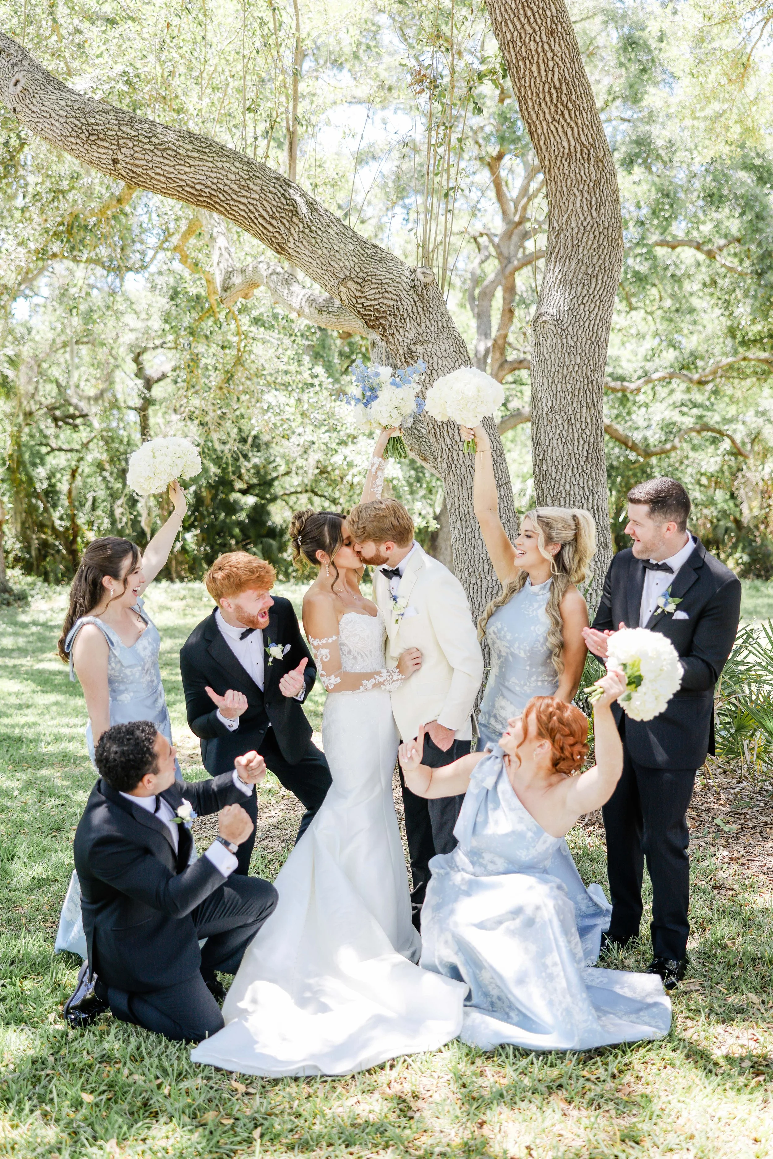 A wedding ceremony outdoors under trees, with the bride and groom kissing. Surrounded by friends and family, some are kneeling, clapping, and holding bouquets of blue and white flowers. The bride wears a white gown, and the groom is in a white tuxedo. The scene is sunny and lively.
