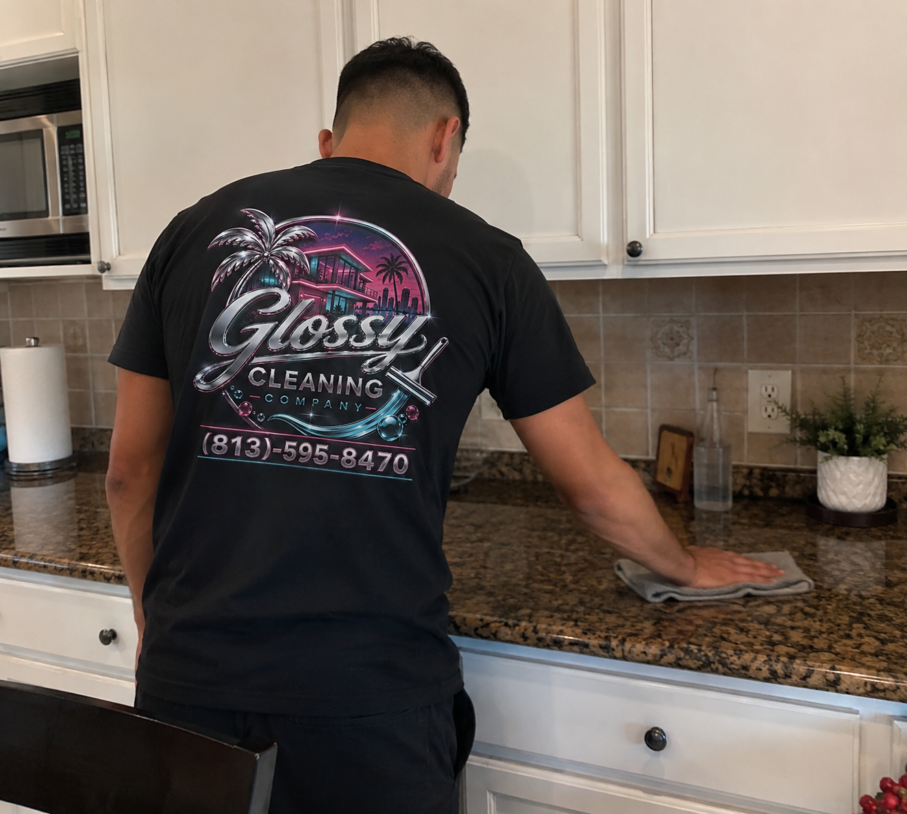A man in a black T-shirt cleaning a kitchen countertop with a cloth. The T-shirt has a logo for Glossy Cleaning Company with palm trees, a building, and a phone number.
