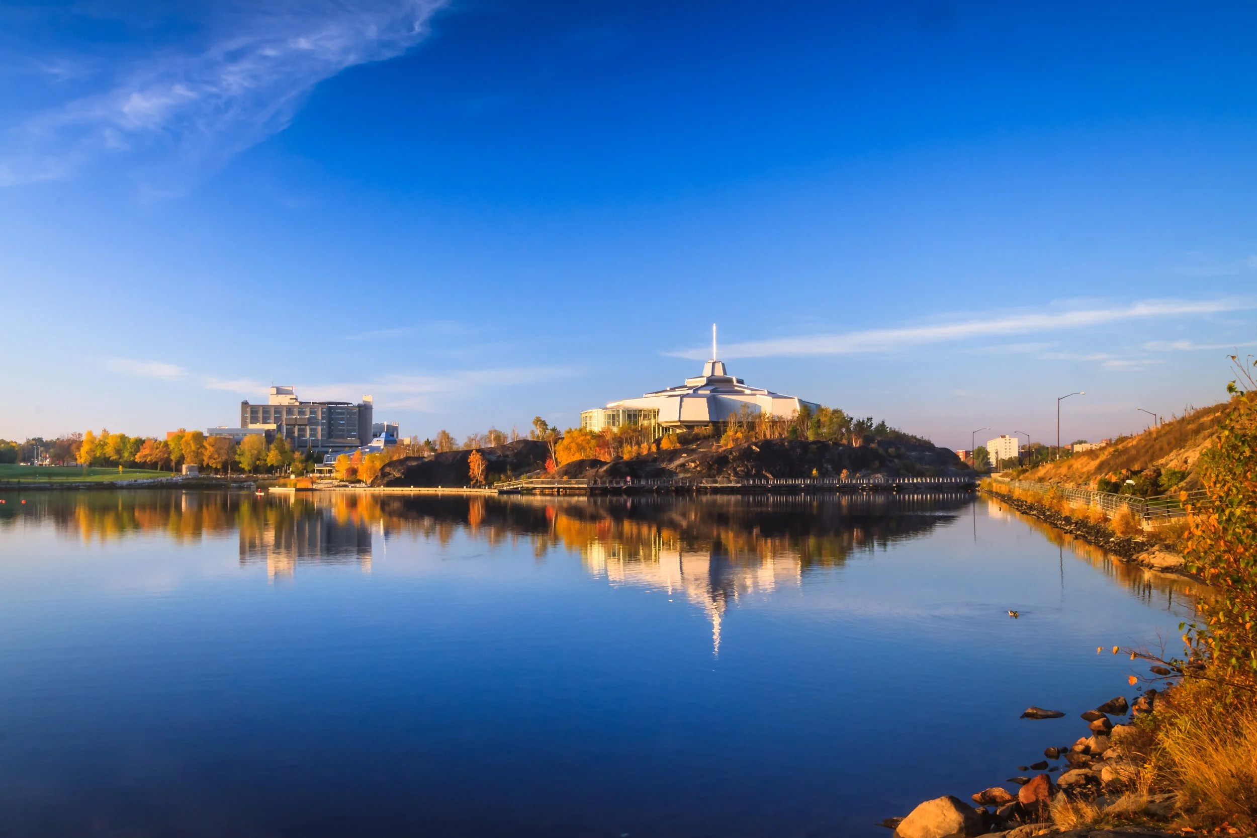 City skyline with modern building shaped like a spaceship, reflected on a river, with autumn trees and a clear blue sky. Showing Science North and Ramsey Lake in Sudbury Ontario.