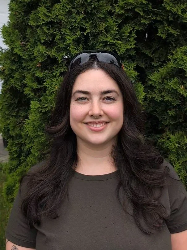 A woman with long dark hair and sunglasses on her head, smiling in front of a green leafy background. Ashleigh Gauthier, home inspector for Spotlight Home Inspections.