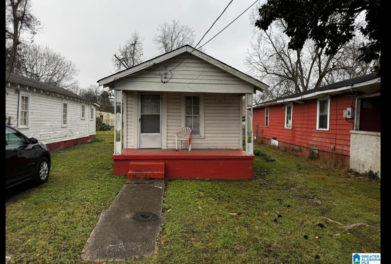 Front view of a small house with a porch, a plastic chair, and a concrete walkway, surrounded by other houses and trees.