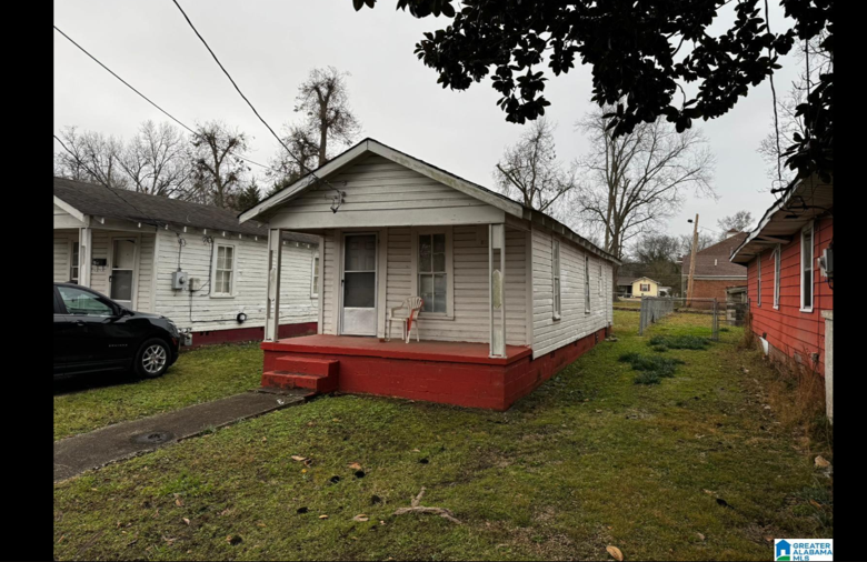 Front view of a small, single-story house with white horizontal siding and a red foundation and steps, a small porch with a chair, and a grassy yard. There are neighboring houses and trees in the background, and an overcast sky.