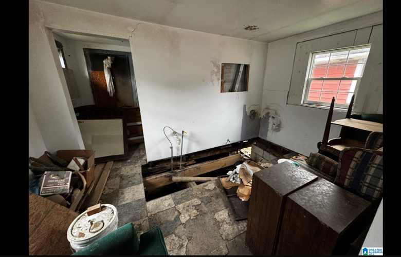 Room with a large hole in the floor, exposed wooden beams, and damaged walls. Furniture and clutter are scattered around.