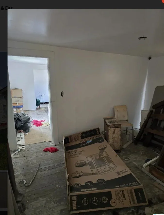 A room under renovation with cardboard boxes, a chair, and scattered debris; an adjoining room with more clutter and furniture.
