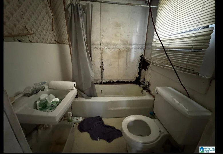 Bathroom with damaged wall, open bathtub, sink with cleaning supplies, toilet, and a window with blinds.