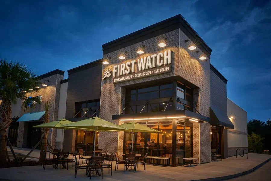 Exterior of a bakery restaurant named 'First Watch' during evening, with outdoor seating, green umbrellas, and illuminated sign.