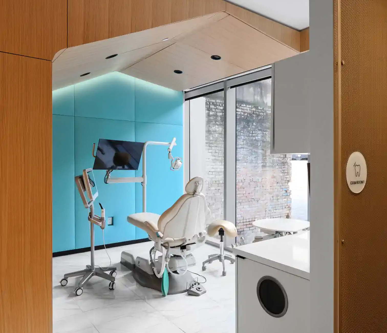 A modern dental examination room with a dental chair, overhead light, monitor, and various dental tools, located near large windows with a brick wall outside.