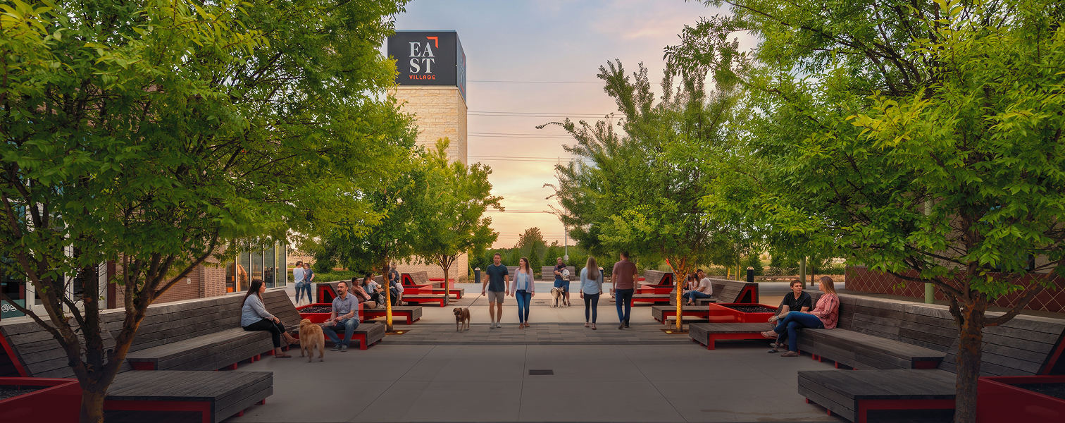 People sitting and walking in an outdoor plaza with trees, benches, and a modern building at sunset.