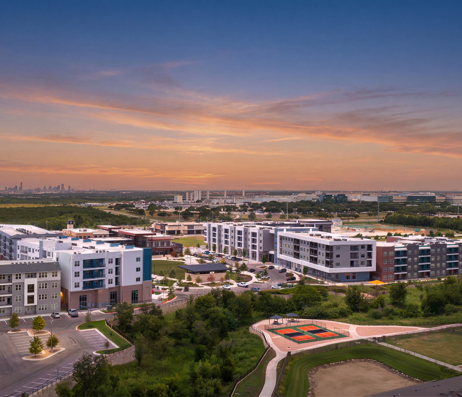 Aerial view of modern apartment complex with parking lots, tennis courts, and green spaces at sunset, with a city skyline in the distance.