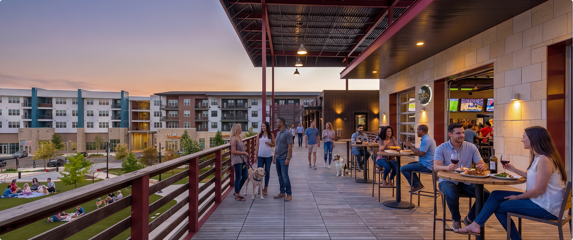 People enjoying drinks and food at an outdoor rooftop bar in an apartment complex during sunset, with some walking dogs and others seated indoors, overlooking a landscaped park.