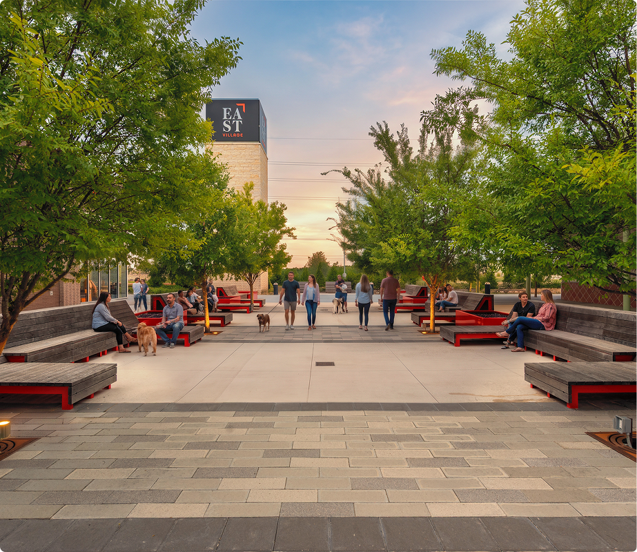 People sitting on benches and walking in a landscaped outdoor area with trees, during sunset, in East Village, with a building labeled 'EAST' in the background.