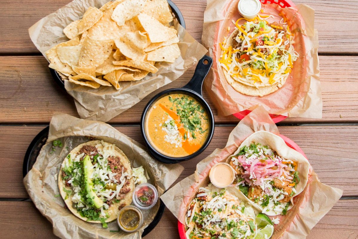 A top-down view of a wooden table with Mexican food including nachos, three tacos, and a bowl of soup.