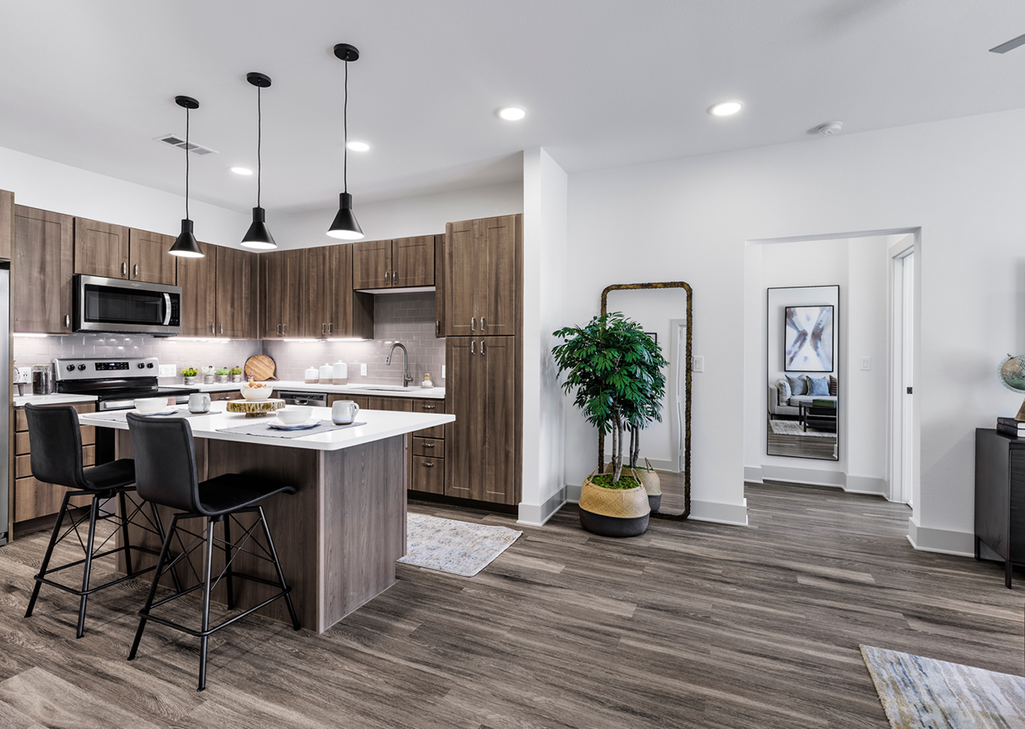 Modern kitchen with wooden cabinets, white countertops, black pendant lights, and a kitchen island set for breakfast with dishes and cups. There is a large green potted plant next to a mirror and a hallway leading to another room with artwork and a classical globe on a dark furniture piece.