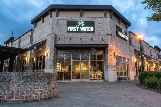 Exterior view of a shopping center with a store called First Watch, featuring brick walls, large glass windows, and outdoor lighting at dusk.