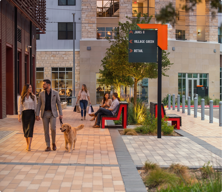 People walking and sitting on benches in an urban plaza, with modern buildings in the background, a dog on a leash, and a digital directional sign.
