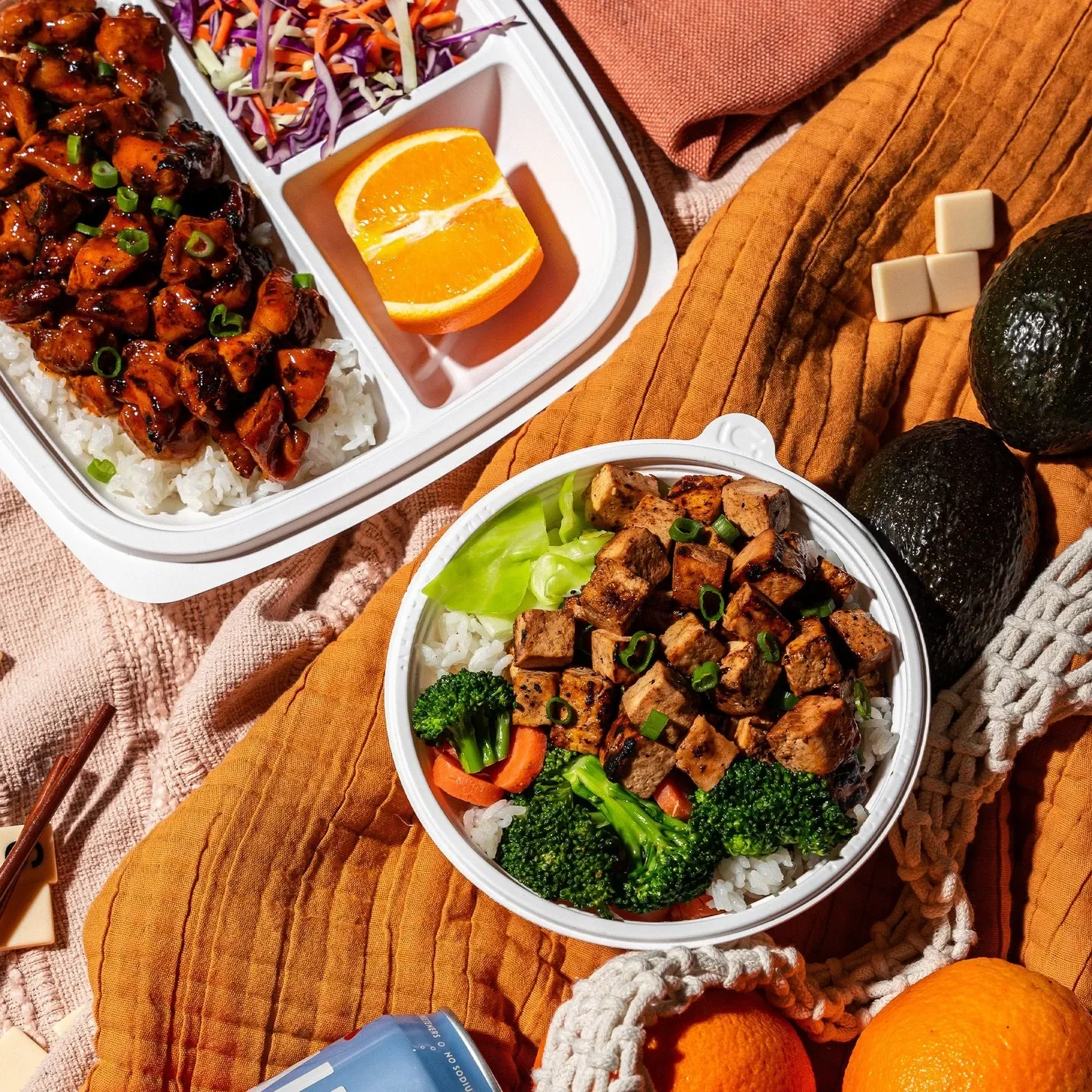 A meal with bowls of rice topped with diced tofu and vegetables, a plate of Asian-style chicken and rice, oranges, and avocados, all set on a textured orange and brown cloth display.