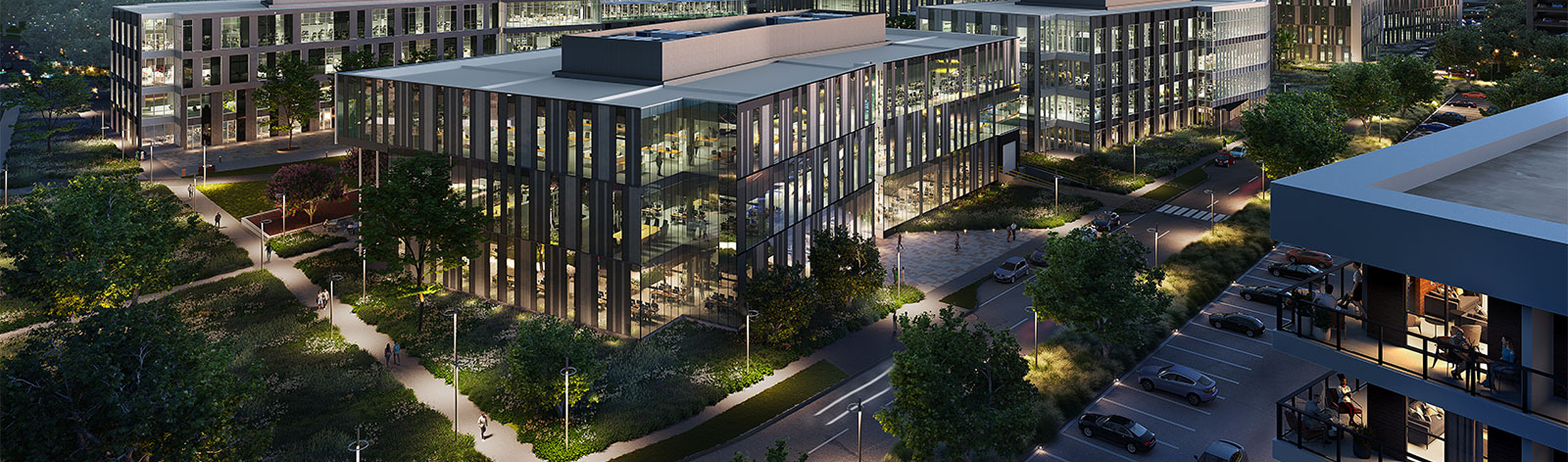 Nighttime view of a modern office building with glass walls, surrounded by trees, illuminated walkways, and parked cars.