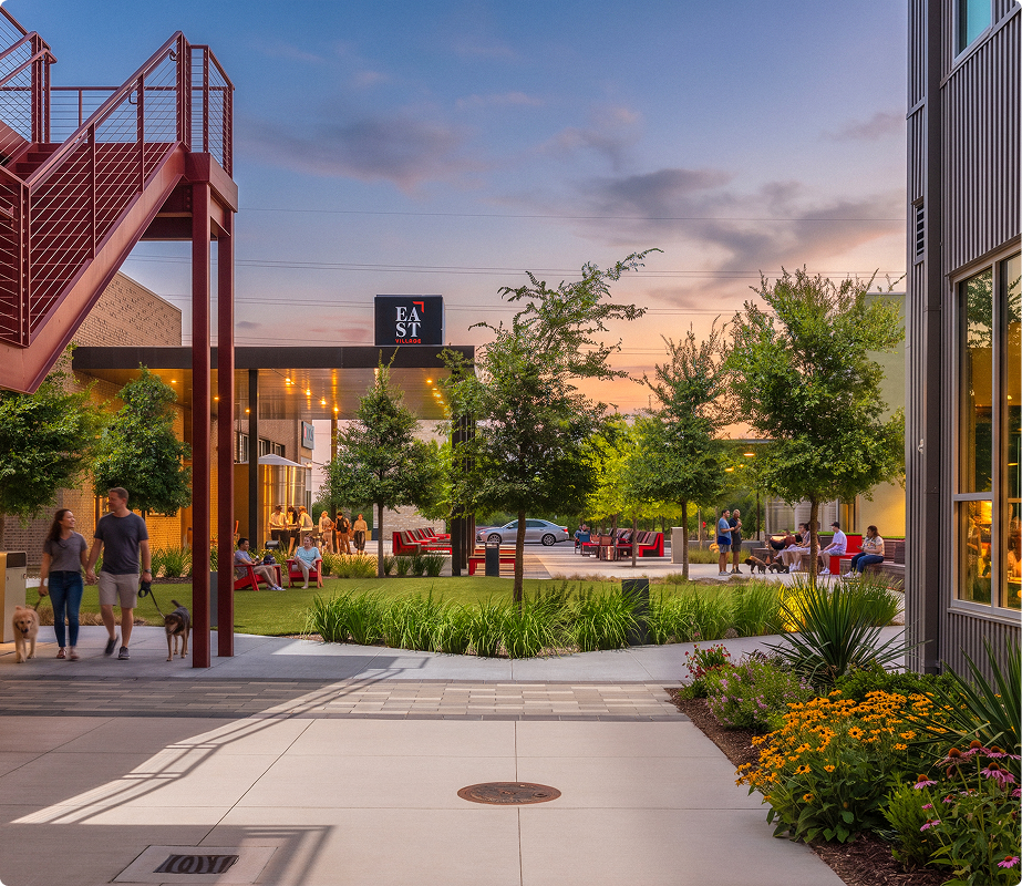 People walking dogs along a modern shopping or dining plaza during sunset, with trees, outdoor seating, and a building with an 'EAST' sign in the background.