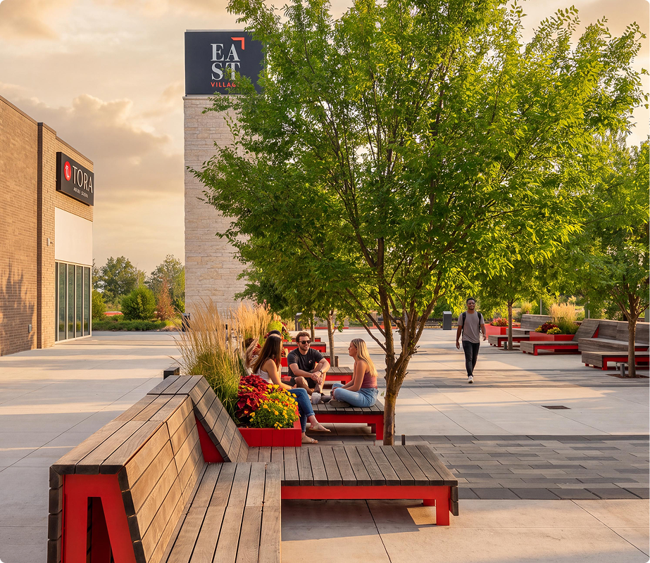 People sitting on benches under trees at an outdoor shopping or dining plaza, with stores and greenery visible in the background during sunset.