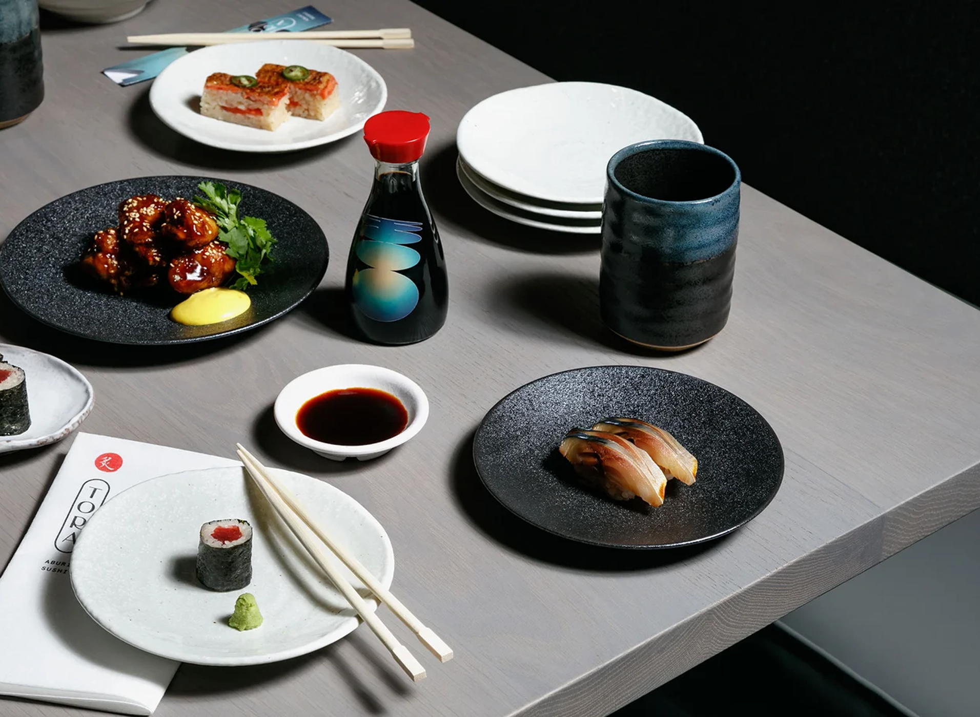 A dining table set with Japanese cuisine, featuring sushi, sashimi, soy sauce, wasabi, and a plate of glazed chicken with cilantro and sesame seeds, along with a soy sauce dish, cups, and stacked white plates.
