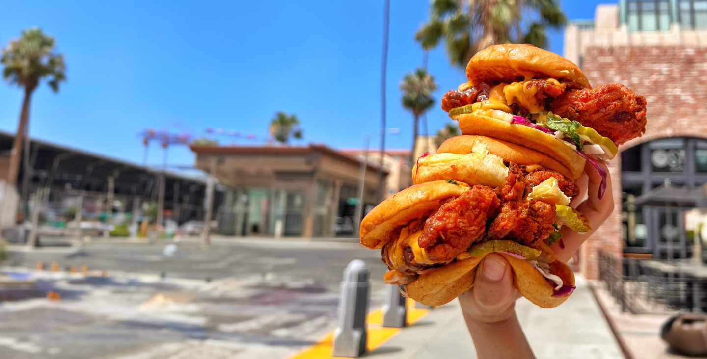 Hand holding a large sandwich with fried chicken, pickles, lettuce, tomato, cheese, and a bun on a city street in bright daylight.