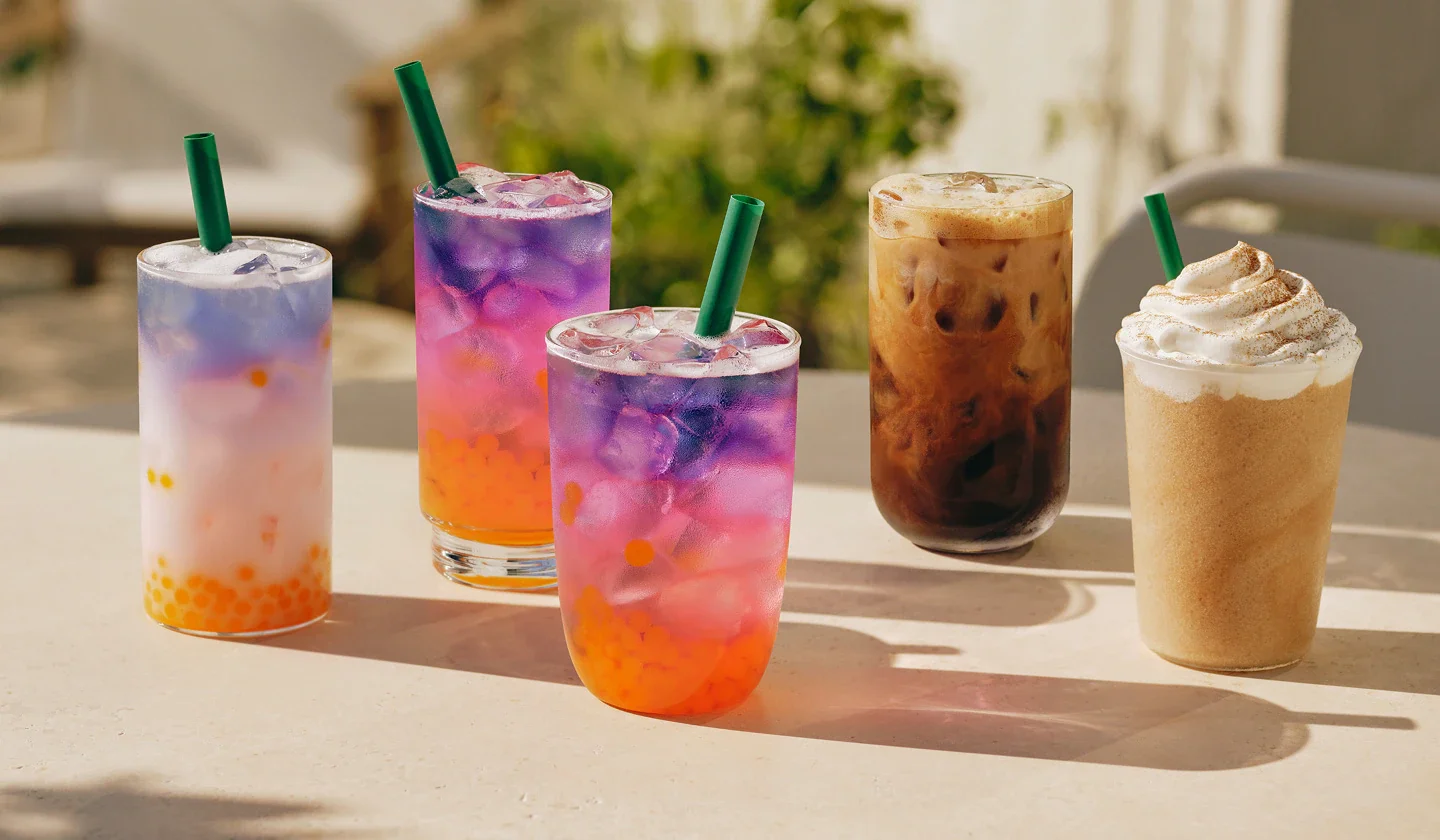 Five colorful iced beverages on a table outdoors, including two purple drinks, a caramel-colored coffee with ice, and a frappuccino with whipped cream.