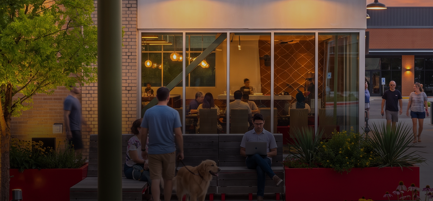 People sitting and walking outside a cafe with large glass windows, some working on laptops and others talking.