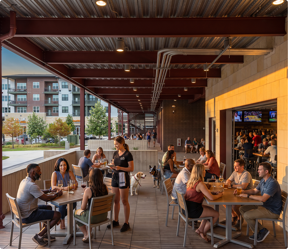 People dining and socializing on a covered outdoor patio of a restaurant during sunset, with view of buildings and trees in the background.
