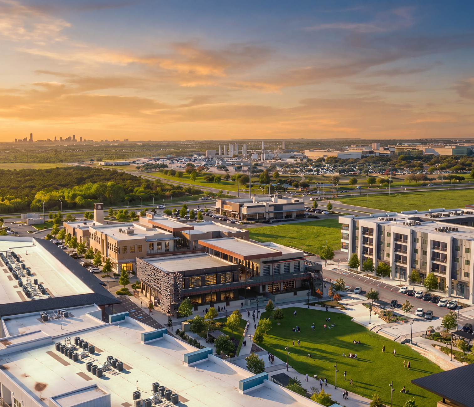 Aerial view of a mixed-use urban area at sunset with buildings, parks, roads, and a city skyline in the distance.