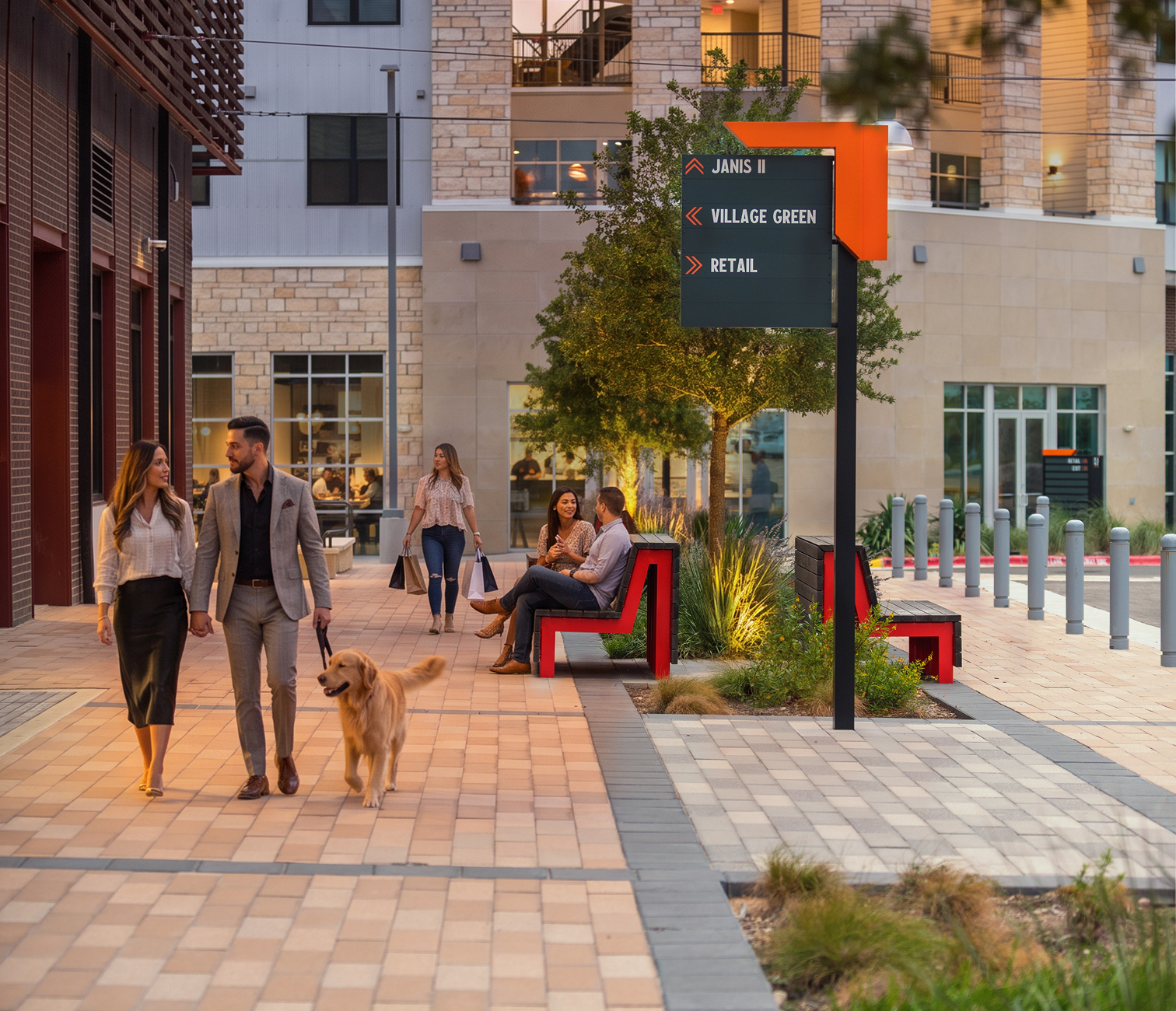 People enjoying a pleasant evening in a modern urban shopping plaza. A couple walks their golden retriever, while two women sit on a bench chatting and another woman walks with shopping bags in hand. The area features stylish benches, greenery, and directional signs for nearby locations.