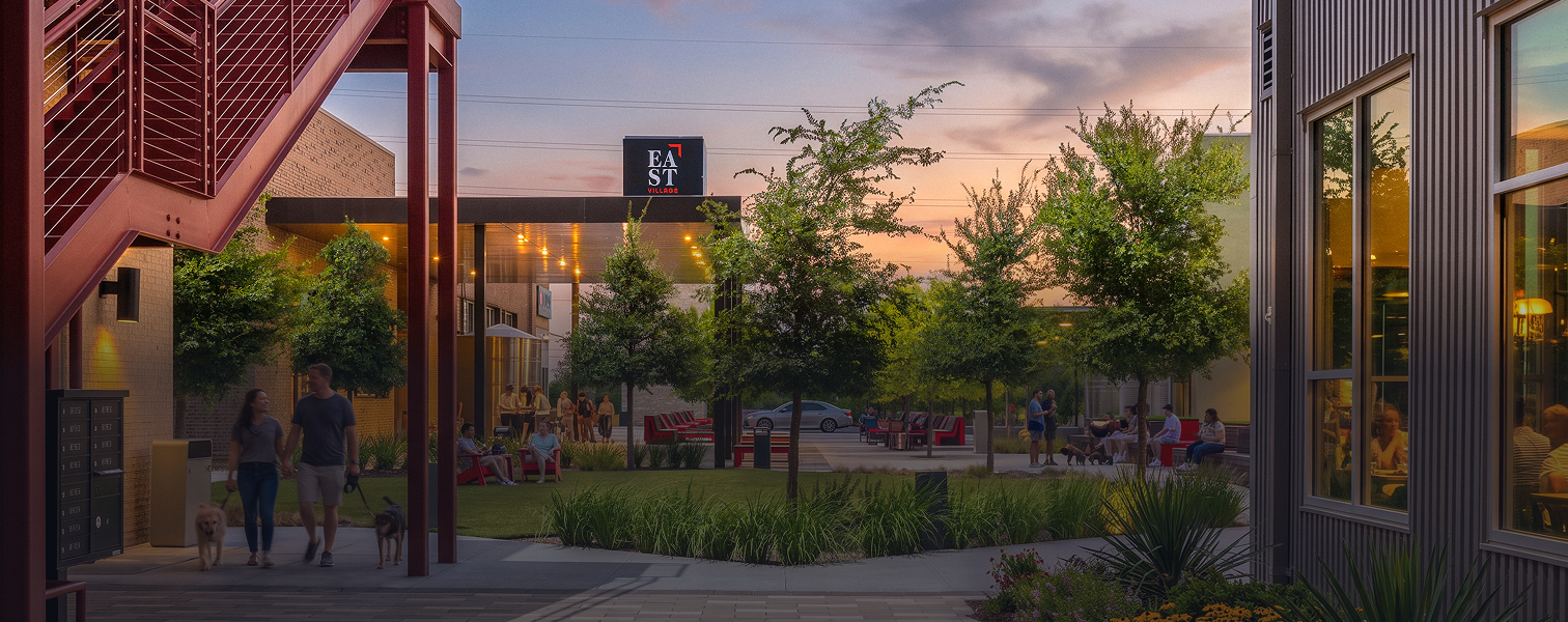 An outdoor scene at dusk with people sitting, walking dogs, and socializing in a landscaped area with trees, benches, and modern buildings, including a restaurant with large windows.
