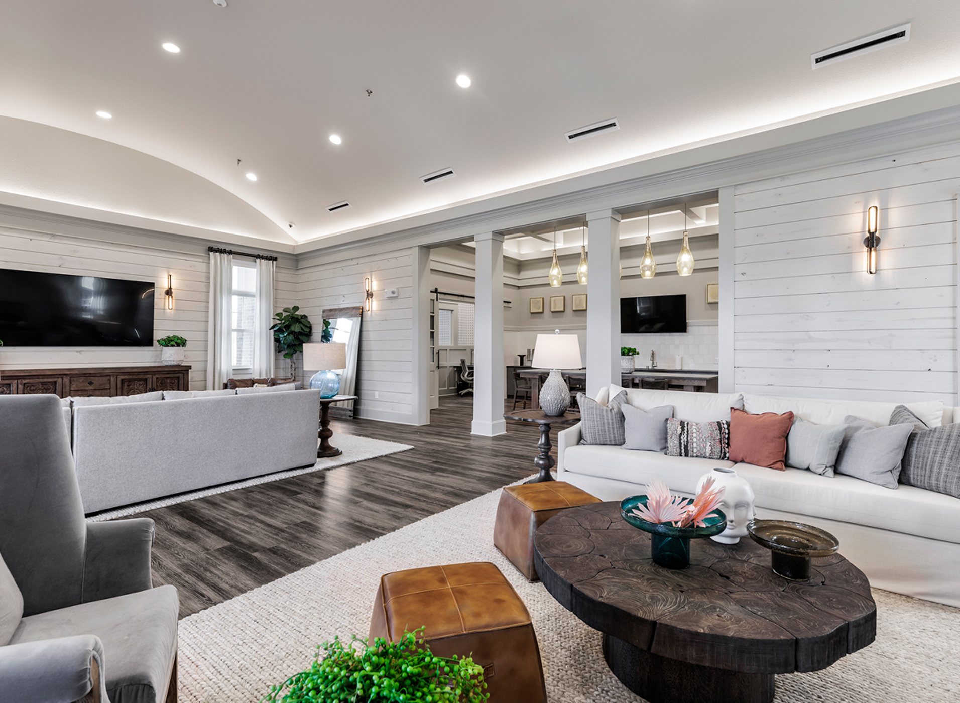 Modern living room with white walls, dark wood flooring, sitting area with grey and white couches, a large flat-screen TV, decorative lamps, pillows, coffee table with decor, and a dining area in the background.